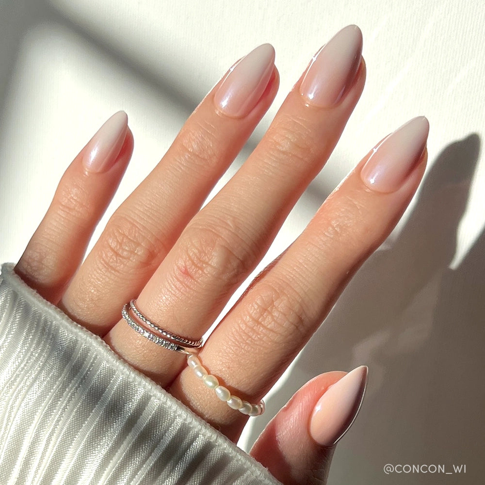 A hand with long, almond-shaped ombré nails in the shade "Fiancéd" by glamnetic, wearing two silver rings and a pearl bead ring. The person is in a white ribbed sweater, photographed under natural sunlight.